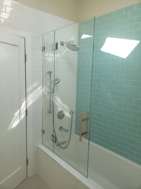 Frameless hinged glass panel over a bathtub with teal tile and wood pull handles